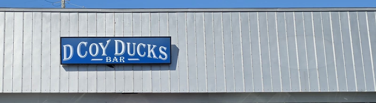 The blue D Coy Ducks Bar sign on a silver metal siding band above the storefront