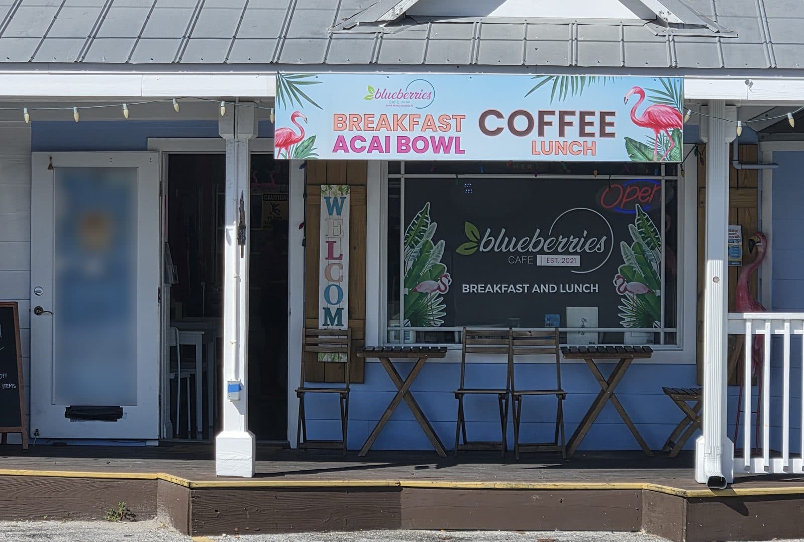 The Blueberries Cafe sign, Breakfast, Acai Bowl, Coffee, Lunch, with flamingos over a blue porch with small bistro tables