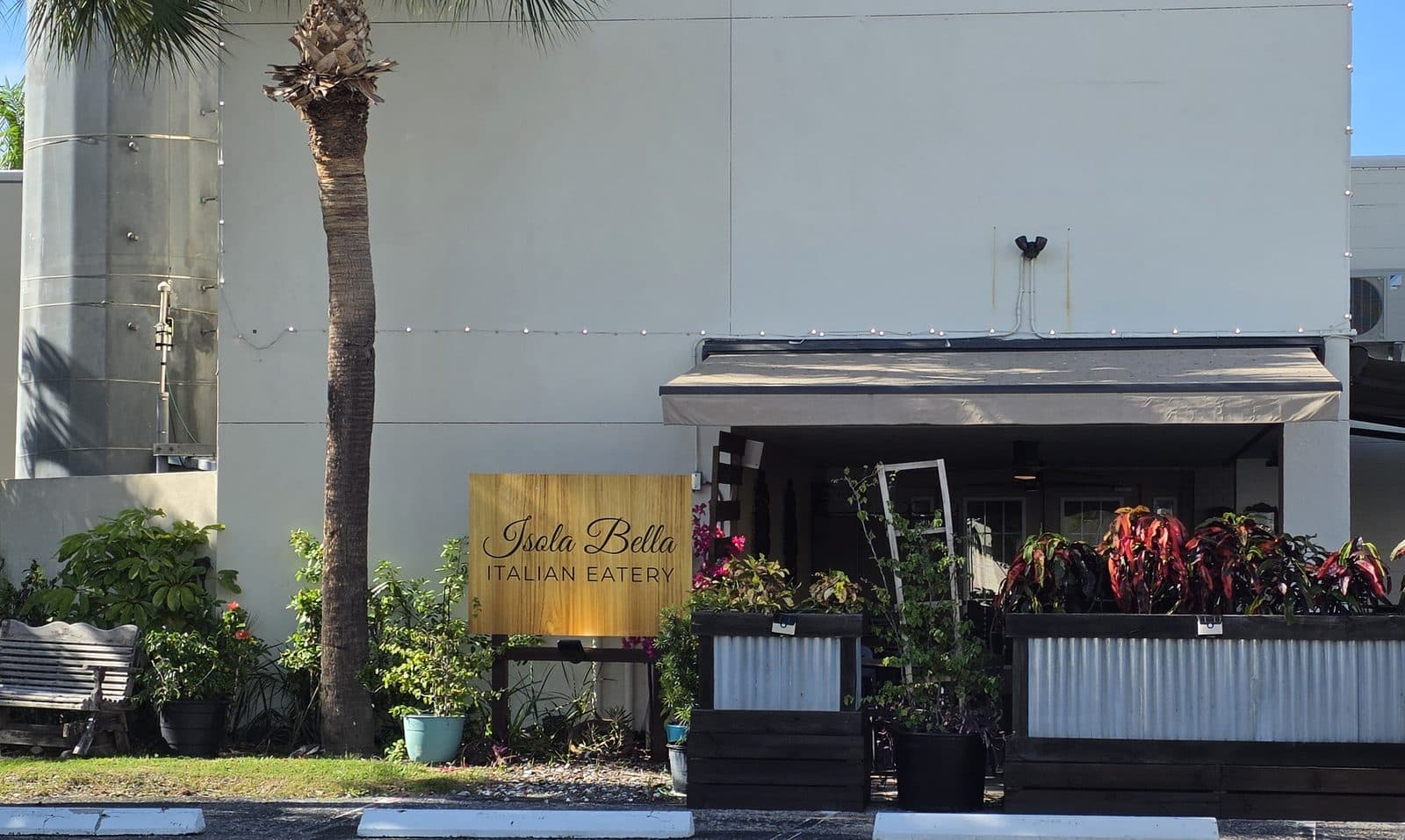 The wooden Isola Bella Italian Eatery sign beside a palm, with a covered patio and red leafed planters behind